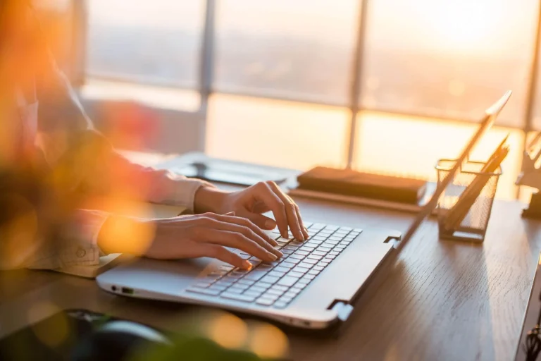 A woman creating website content by typing on a laptop in front of a window.