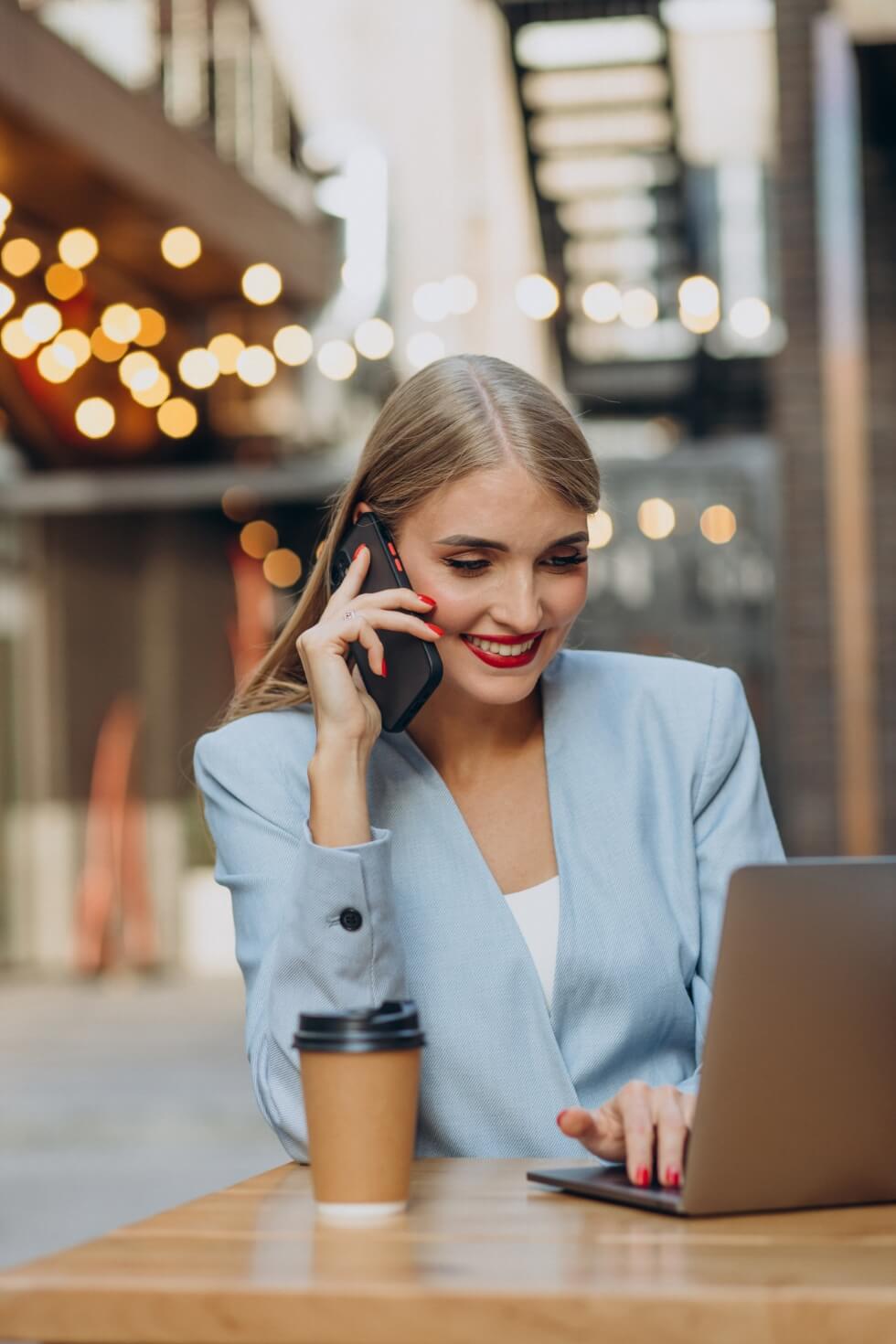 A woman is talking on the phone while sitting at a table with a laptop and coffee.